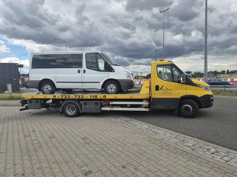 Laweta pomocy drogowej Tuszyn przewozi bus Ford Transit przez parking w pochmurny dzień