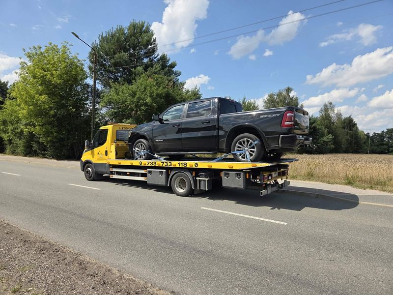 Laweta pomocy drogowej Tuszyn z czarnym pickupem RAM na platformie, na drodze wśród drzew