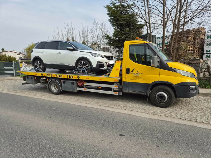 Laweta wiezie okazały, sprawny SUV rodzinny ulicą miasta obok składu palet drewnianych