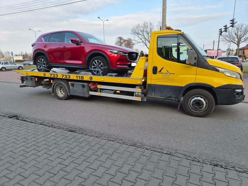 Pomoc drogowa Tuszyn holuje lawetą nieuszkodzonego SUV-a przez skrzyżowanie w mieście w pochmurny dzień