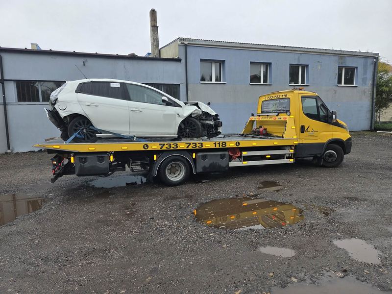 Pomoc drogowa Tuszyn przewozi lawetą mocno uszkodzony po zderzeniu hatchback obok budynku przemysłowego