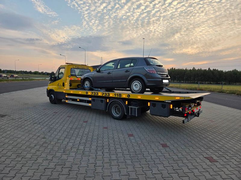 Laweta pomocy drogowej Tuszyn z załadowanym Oplem Astra na parkingu przy drodze ekspresowej o zachodzie słońca