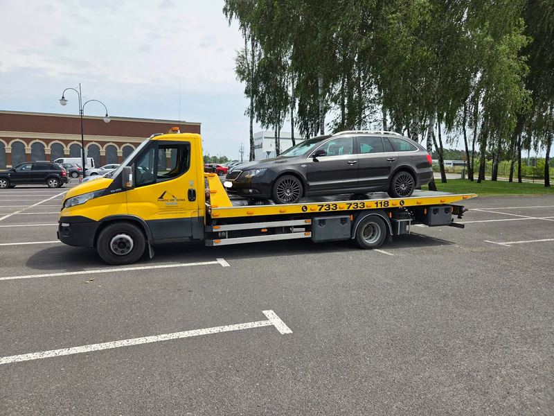 Laweta pomocy drogowej Tuszyn przewozi sprawne kombi przez miejski parking w pochmurny dzień