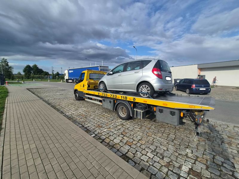 Laweta wiezie mały hatchback przez parking pod ciemnymi chmurami, pomoc drogowa Tuszyn
