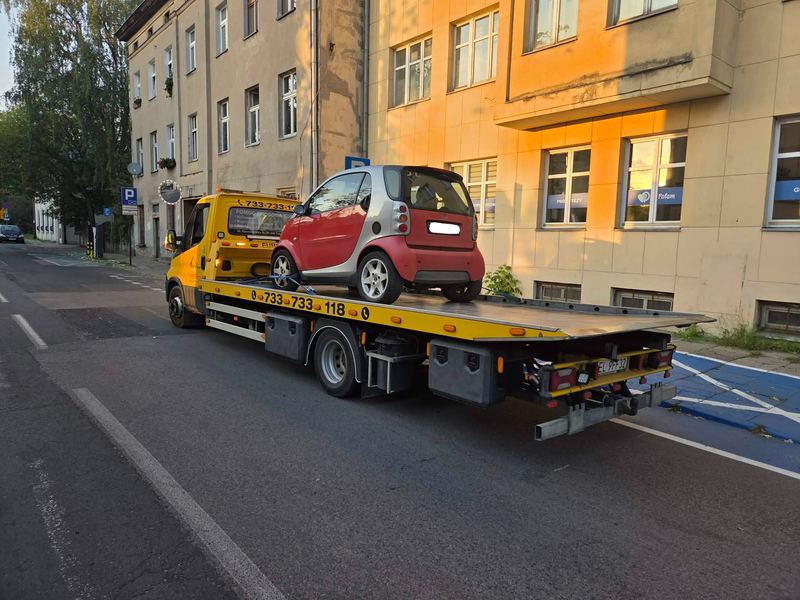 Laweta pomocy drogowej Tuszyn transportuje małe miejskie auto ulicą w centrum miasta